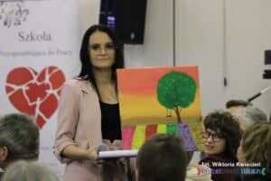 Brunetka prezentująca licytowany obraz z namalowanym drzewem i pejzażem kolorowych pól