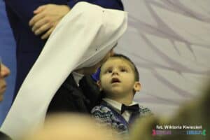 Siostra zakonna w białym welonie trzyma na rękach chłopca, który intensywnie wpatruje się w Jej twarz