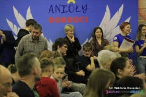 Publiczność wydarzenia. Część osób siedzi, część stoi oparta o ciemnoniebieski baner z napisem "Anioły Dobroci" i namalowanymi wielkimi, białymi skrzydłami