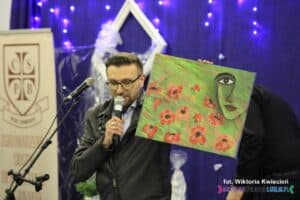 Mężczyzna z mikrofonem prezentuje licytowany obraz w kolorze zieleni, przedstawiający fragment twarzy umieszczonej w prawy górnym rogu., pozostałą część malowidła wypełniają czerwone kwiaty z czarnymi środkami