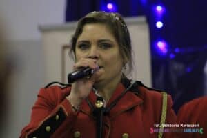 Kobieta z mikrofonem, ubrana w czerwoną marynarkę od munduru Orkiestry Dętej Gminy Końskowola