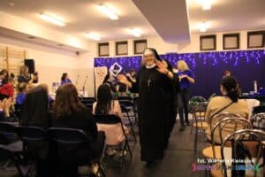 pomiędzy rzędami krzeseł na widowni utworzono taneczny "wąż"; na przedzie siostra zakonna, patrzy i uśmiecha się do fotografującego