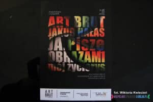 Ulotka promująca wystawę "Art Brut Jakub Hałas. JA. Piszę obrazami mój życiorys" umieszczona na czarnym tle za szybą