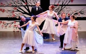 Sześciu tancerzy występuje na scenie w formalnych strojach. Kobiety noszą kremowe sukienki, a mężczyźni czarne garnitury. W tle znajduje się ściana w kwiatowe wzory