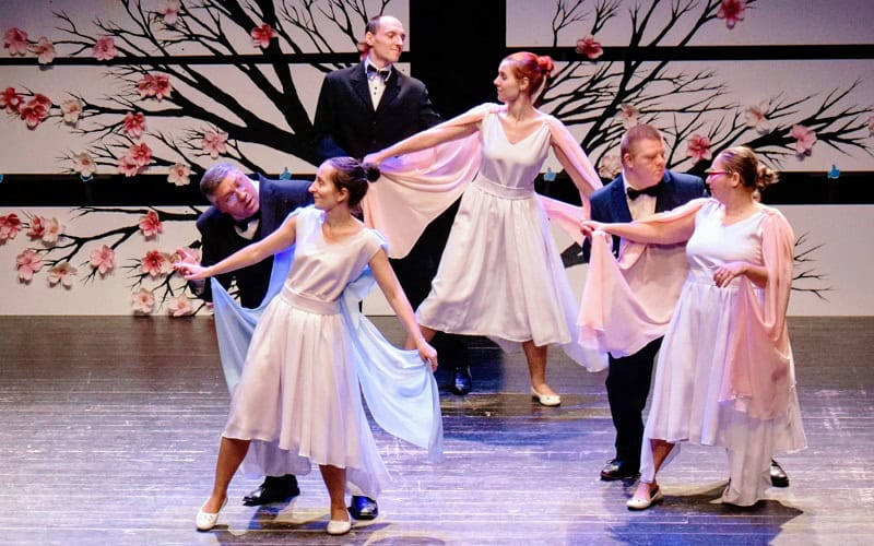 Sześciu tancerzy występuje na scenie w formalnych strojach. Kobiety noszą kremowe sukienki, a mężczyźni czarne garnitury. W tle znajduje się ściana w kwiatowe wzory