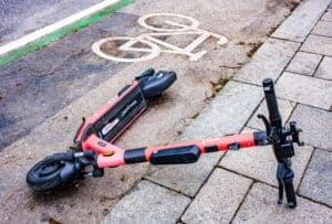 Czerwona hulajnoga elektryczna leży na boku w pobliżu oznakowania ścieżki rowerowej na betonowym chodniku.