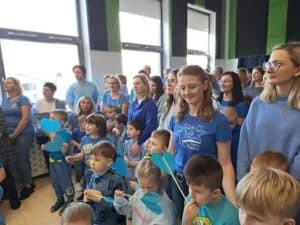 Grupa dorosłych i dzieci, ubranych głównie na niebiesko, stoi w sali. Dzieci trzymają na patykach niebieskie wycinanki w kształcie serc.