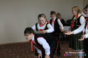 Dziewczynka inscenizuje wers piosenki: "I tym wałkiem, kiedy chciała Swego męża krasnoludka wałkowała"; trzyma drewniany wałek do ciasta na plecach schylonego przed nią chłopca