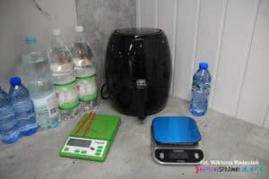 Na blacie kuchennym w rogu ustawiono air fryer, wagi elektroniczne oraz butelki z wodą