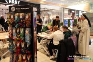 Stojak ze "Skarpetkami PoMocy"; w tle przy stole uczestnicy warsztatów ze Stowarzyszenia „Źródło” Osób Niepełnosprawnych Umysłowo i ich Rodzin i Przyjaciół