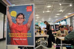 Stojak z plakatem Skende z napisem: '21-22 marca. Świętujemy Światowy Dzień Zespołu Downa. Dołącz do nas!' Na plakacie widnieje zdjęcie młodego mężczyzny w czerwonej koszulce i słuchawkach na uszach, który ma uniesione do góry rozłożone ręce