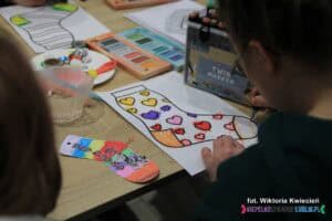 Dziecko przy stole koloruje flamastrami kartkę z konturem skarpetki w serduszka. Obok po lewej stronie leży ukończona kolorowa zakładka w kształcie skarpetki z wizerunkiem zebry. Na blacie wokół leżą materiały plastyczne, m.in. farby i flamastry