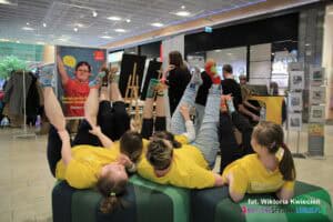 Pasaż handlowy Skende: na zielonych pufach pięć osób w jednakowych żółtych koszulkach leży na plecach z uniesionymi nogami, prezentując założone na stopy kolorowe skarpetki nie do pary