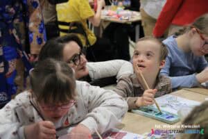 Pani mgr Iwona Lal, Dyrektor SOSW nr 2 w Lublinie, kuca pomiędzy dwojgiem malujących dzieci – chłopcem i dziewczynką. Z uśmiechem patrzy na chłopca, który z lekko uniesioną głową i subtelnym uśmiechem spogląda w górę