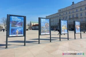 Plac Litewski w centrum Lublina skąpany w promieniach słońca, a w tle majestatyczny budynek Poczty Polskiej. Na placu stoją cztery wystawowe stojaki, na których umieszczono plakaty z napisem 'Różni, ale równi - historie pełne kolorów'. Każdy plakat przedstawia kolaż zdjęć oraz krótki opis aktywności, tworząc kolorową, inspirującą prezentację