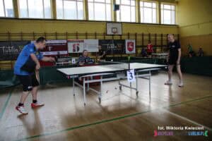 Dwóch zawodników rywalizuje w meczu tenisa stołowego przy stole nr 3. Zawodnik po lewej stronie, ubrany w niebieską koszulkę, wykonuje serwis. Zawodnik po prawej, w czarnej koszulce jest przygotowany do odbicia piłeczki