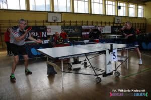 Dwóch zawodników w trakcie dynamicznej wymiany piłeczki w meczu tenisa stołowego przy stole nr 5. Zawodnik po lewej jest w trakcie odbicia piłeczki, a drugi - po prawej stronie stołu (w czarnej koszulce) przygotowuje się do przyjęcia. W tle widać sędziego