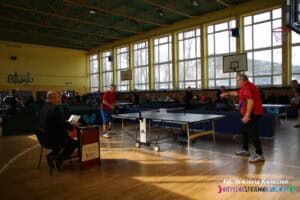 Widok na salę gimnastyczną, w której odbywają się rozgrywki w tenisa stołowego. Na pierwszym planie dwóch zawodników w czerwonych koszulkach rozgrywa mecz