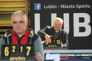 Dwóch sędziów siedzących przy dwóch osobnych stołach. Na pierwszym planie, po lewej stronie, widać rozmazaną postać, ostrość ustawiono na sędziego z tyłu, który przekręca wyniki na tablicy punktowej (wynik to 12-10 w pierwszym secie, a stan meczowy to 0-1)