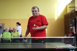 Zawodnik z WTZ Nałęczów w czerwonej koszulce trzyma prawą dłoń z rozłożonymi palcami do góry na wysokości swojego brzucha. Nad jego dłonią widoczna jest biała piłeczka uchwycona w ruchu