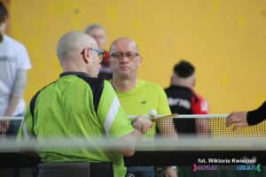 Na zdjęciu widać dwóch zawodników w okularach, siedzących na wózkach, ubranych w prawie identyczne neonowe koszulki. Na pierwszym planie widać plecy jednego z nich, który trzyma rakietkę w prawej dłoni. Na rakietce spoczywa biała piłka do tenisa stołowego