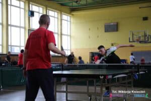 Młody mężczyzna w trójkolorowej koszulce stoi przy stole tenisowym w dynamicznej pozie. Jest pochylony do przodu, stoi na jednej nodze, a drugą unosi w tył. Zawodnik odbija piłeczkę, koncentrując się na akcji. Jego przeciwnikiem jest mężczyzna stojący tyłem do fotografującego, ubrany w czerwoną koszulkę z napisem 'WTZ NAŁĘCZÓW' na plecach