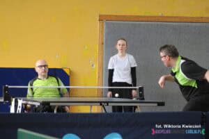 Dwóch mężczyzn rozgrywa mecz. Na końcu stołu (przodem do fotografującego) siedzi mężczyzna w neonowej koszulce na wózku. Jego przeciwnikiem jest mężczyzna stojący w dynamicznej pozycji. Tuż nad stołem przy siatce unosi się biała piłeczka, a w tle widać młodą wolontariuszkę, która asystuje przy rozgrywce