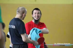 Na zdjęciu stoją dwaj zawodnicy. Mężczyzna stojący przodem do fotografującego to brunet z brodą, ubrany w czerwoną koszulkę, trzyma niebieski ręcznik i szeroko uśmiecha się, patrząc na stojącego obok mężczyznę w granatowej koszulce z białym napisem „Aleksander” na plecach, który trzyma rakietkę w prawej ręce