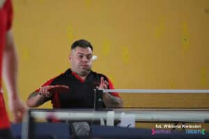 Mężczyzna na wózku trzyma rakietkę w prawej ręce, zagrywając piłeczkę. Jego lewy policzek jest wydęty, jakby wypuszczał powietrze, a piłeczka znajduje się w locie na wysokości jego oczu