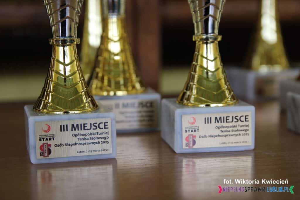 Zbliżenie na metalową tabliczkę przytwierdzoną do podstawy pucharu. Tabliczka zawiera inskrypcję: "III miejsce, Ogólnopolski Turniej Tenisa Stołowego Osób Niepełnosprawnych 2025, Lublin 21-23 marca 2025 r."
