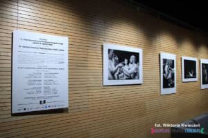 Na tle drewnianej ściany, wisi ekspozycja fotografii. W centrum znajduje się plansza opisowa, a obok wiszą cztery czarno-białe fotografie