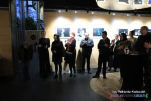 Uczestnicy wernisażu ustawili się w rzędzie pod ścianą, na której wiszą bardzo jasno oświetlone fotografie. Pierwszy mężczyzna od lewej kłania się, a pozostali zgromadzeni biją brawo