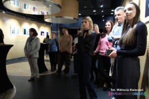 Wei foyer stoi spora grupa osób, zasłuchanych i patrzących przed siebie, poza zasięg fotografującego. Ich uwagę przyciąga coś poza kadrem, tworząc atmosferę skupienia i refleksji