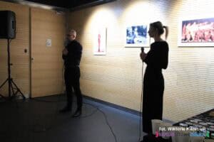 Od lewej stoją ubrani na czarno i pogrążeni w cieniu: Pan Mariusz Kuszpa i Pani Natalia Dudek. Ich ubiór kontrastuje z bardzo jasno oświetloną ścianą za nimi, na której wiszą fotografie, podkreślając różnicę między postaciami a tłem