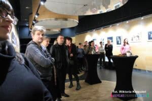 Grupa osób stojących w lekkim półkolu. Na środku widoczne stoliki obleczone na czarno, na których znajdują się woda i przekąski. Na ścianach w tle wiszą fotografie, dobrze oświetlone reflektorami