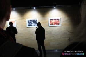 Na środku stoi ubrany na czarno i pogrążony w cieniu Pan Robert Pranagal. Jego ubiór kontrastuje z bardzo jasno oświetloną ścianą za nim, na której wiszą fotografie, podkreślając różnicę między postacią a tłem