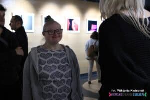 Dziewczynka w okularach. Wokół niej stoją inni ludzie, ale tylko ona patrzy prosto w obiektyw aparatu i uśmiecha się