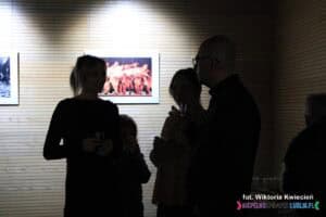 Mocno ocienione sylwetki trzech dorosłych osób i jednego dziecka, każdy trzyma szklankę z napojem. Tło to jasno oświetlona ściana, na której wiszą fotografie