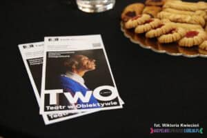 Na czarnym tle leżą dwie ulotki 16. edycji Międzynarodowego Konkursu Fotograficznego TwO, w w rogu fragment złotej tacy z ciastkami