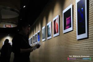 Ocieniona sylwetka mężczyzny stojącego twarzą do ściany z mocno oświetlonymi fotografiami, skupionego na trzymanej w rękach ulotce