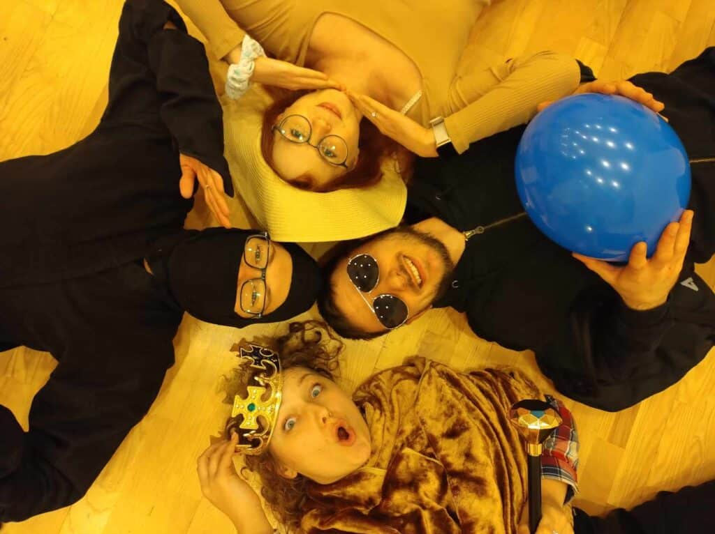 Na podłodze, stykając się głowami leżą cztery osoby; każda ma inne przebranie (królowa, ninja, dama w kapeluszu, mężczyzna w ciemnych okularach)