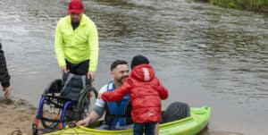 Mężczyzna w kajaku, przy brzegu rzeki, utrzymuje kontakt wzrokowy z dzieckiem w czerwonej kurtce, które jest odwrócone tyłem do aparatu. Obok niego stoi drugi mężczyzna, trzymając wózek inwalidzki znajdujący się na piaszczystym brzegu.