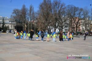 Grupa przedszkolna pod opieką pedagogów, stojąca w okręgu na Placu Litewskim w Lublinie. Dzieci mają na sobie odblaskowe kamizelki oraz niebieskie elementy garderoby i niebieskie balony