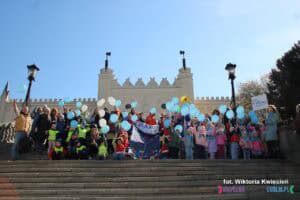 Grupa osób z przewagą dzieci w wieku przedszkolnych stojąca na schodach prowadzących do Zamku Lubelskiego. Dzieci trzymają niebieskie i białe balony oraz transparenty wspierające autyzm