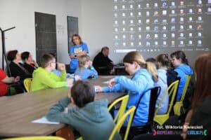 Grupa dzieci i młodzieży zgromadzonej wokół stołu, w tle stoi Pani Sylwia Góźdź, Dyrektor Środowiskowego Domu Samopomocy "Mozaika
