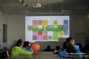 Grupa dzieci przy stole patrzy na ścianę przed sobą, na której wyświetlono slajd z napisem "Piękne Kaczątko" i kolorowym logo ŚDS "Mozaika"