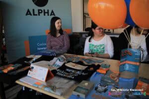 Trzy osoby siedzą przy stoisku Fundacji Alpha; na stoliku ułożono materiały informacyjno-promocyjne