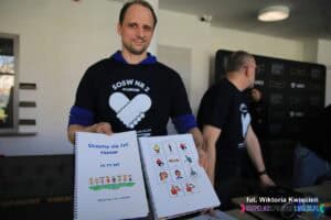 Mężczyzna w koszulce z logo Specjalnego Ośrodka Szkolno-Wychowawczego nr 2 w Lublinie pokazuje fotografującemu książki dydaktyczne do komunikacji alternatywnej pt. "Uczymy się żyć razem. Ja Ty My"