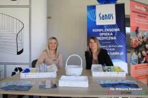 Dwie kobiety przy stoisku Centrum Medycznego Sanus. Na stoliku ułożono materiały informacyjne oraz upominki dla najmłodszych