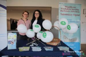 Dwie kobiety na stoisku Niepublicznego Przedszkola Specjalnego „Tajemniczy Ogród”; na stoliku ułożono materiały informacyjne oraz białe balony z logo placówki